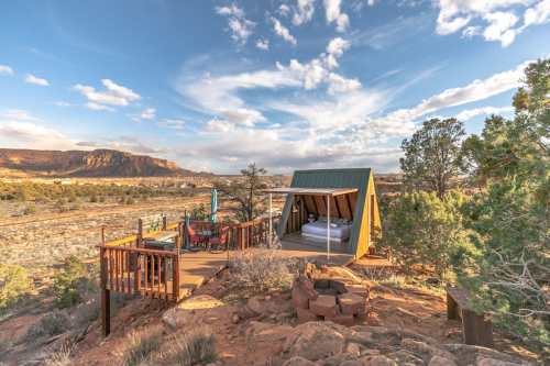 A cozy cabin on a hillside with a deck, surrounded by desert landscape and mountains under a blue sky.