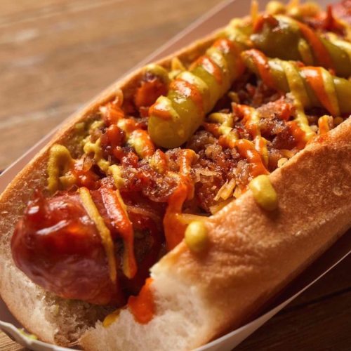 A close-up of a hot dog topped with mustard, ketchup, and onions in a paper tray.