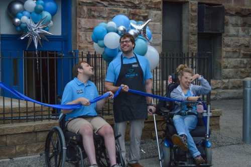 Three individuals, two in wheelchairs, smile while holding a blue ribbon in front of a festive backdrop with balloons.