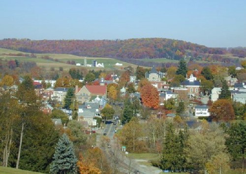 A scenic view of a small town surrounded by autumn-colored trees and rolling hills under a clear blue sky.