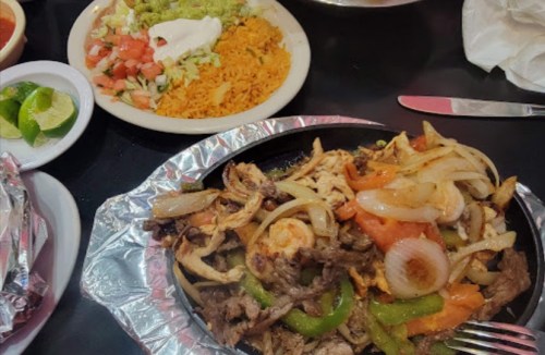 A sizzling platter of mixed grilled meats with onions and peppers, served with rice, salad, and lime wedges.