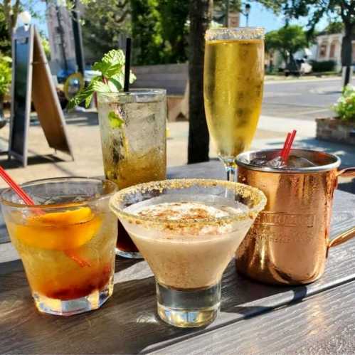 A variety of colorful cocktails on a wooden table, including a martini, a copper mug drink, and a sparkling beverage.