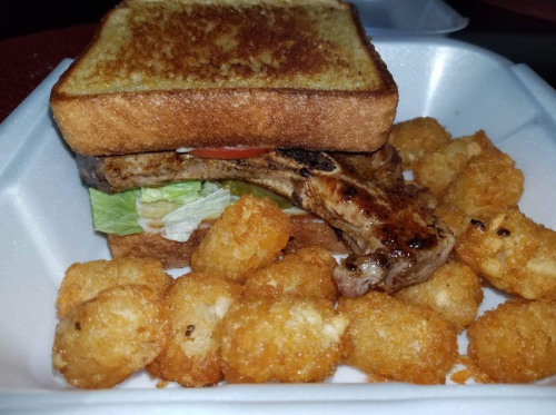 A takeout container with a sandwich featuring grilled meat and lettuce, accompanied by crispy tater tots.