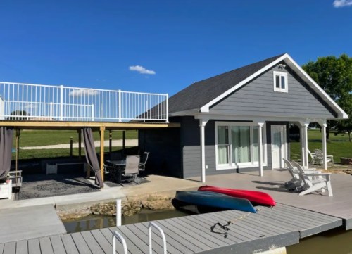 A modern gray house by a lake with a deck, dock, and kayaks, set against a clear blue sky.