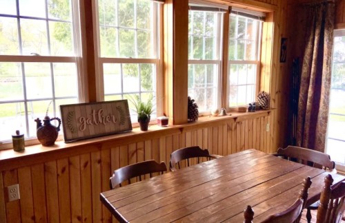 Cozy dining area with a wooden table, chairs, and large windows, featuring a "gather" sign and potted plants.
