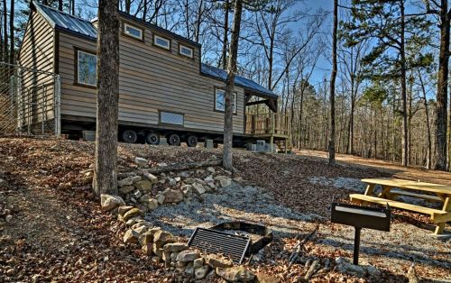A tiny house on wheels surrounded by trees, with a picnic table and fire pit in a wooded area.