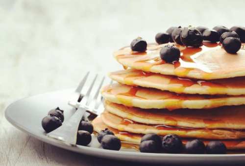 A stack of pancakes drizzled with syrup, topped with fresh blueberries, served on a plate with a fork and knife.