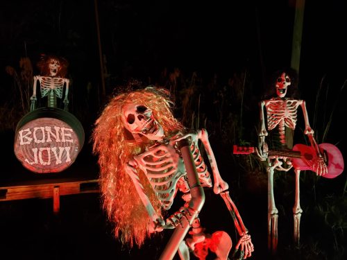 Three skeletons in a dark setting, one with a guitar, another with a sign reading "BONE JOVI," creating a spooky rock band scene.