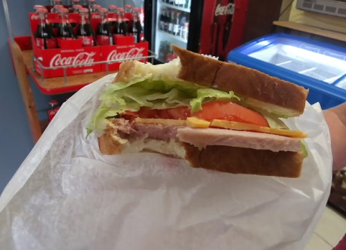 A hand holding a sandwich with lettuce, tomato, and cheese, wrapped in paper, with Coca-Cola bottles in the background.