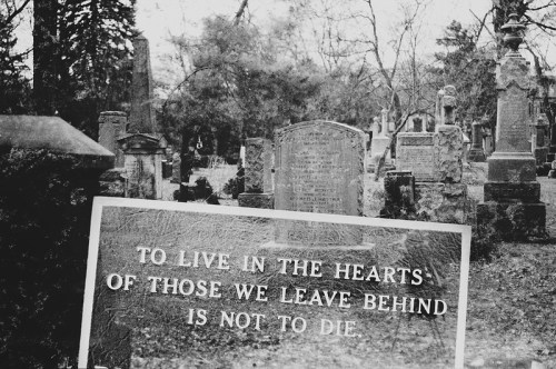 A black-and-white photo of a cemetery with a gravestone and a sign reading, "To live in the hearts of those we leave behind is not to die."