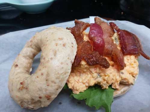A toasted bagel filled with creamy egg salad, topped with crispy bacon and lettuce, served on parchment paper.