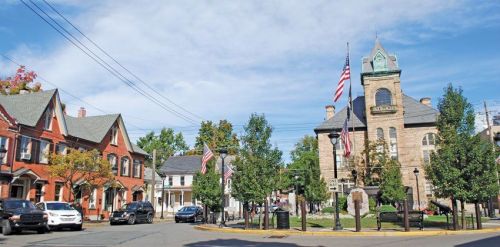 A quaint town square featuring historic buildings, trees, and American flags under a blue sky.