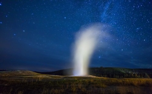 A geyser erupts under a starry night sky, illuminating the landscape with a misty plume.