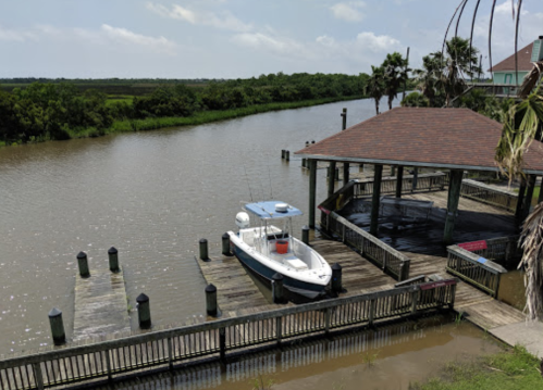 A boat docked at a wooden pier beside a calm river, surrounded by lush greenery and a cloudy sky.
