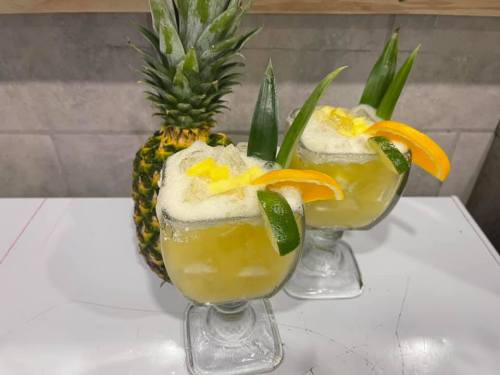Two tropical cocktails with pineapple and orange garnishes, served in glass cups, next to a fresh pineapple.
