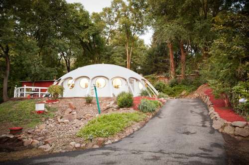 A unique, dome-shaped house surrounded by trees and landscaped gardens, with a gravel driveway leading up to it.