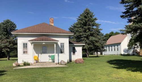 A small white house with a porch and two chairs, surrounded by green grass and trees, with another building in the background.
