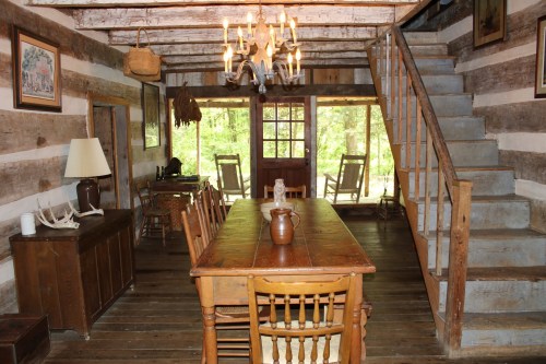 Cozy rustic interior of a log cabin with a wooden dining table, chairs, and a staircase leading to an upper level.
