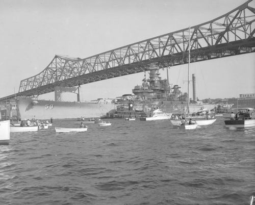 A historic black-and-white photo of a battleship and smaller boats under a large bridge.