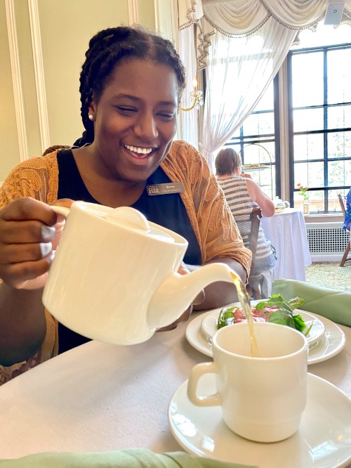 A woman pours tea from a white teapot into a cup, smiling at a dining table with a salad nearby.