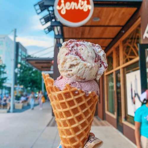 A hand holds a waffle cone filled with pink and white ice cream outside a shop with a sign that reads "Jeni's."