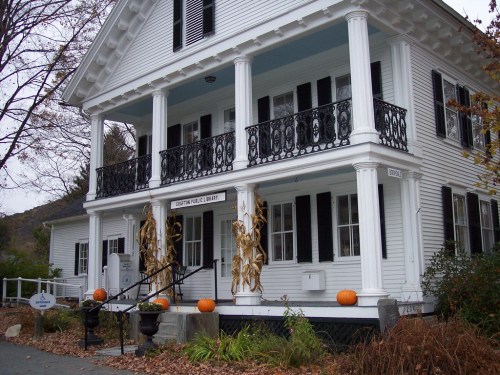 A white, two-story library with black shutters, decorative corn stalks, and pumpkins on the steps, surrounded by autumn foliage.
