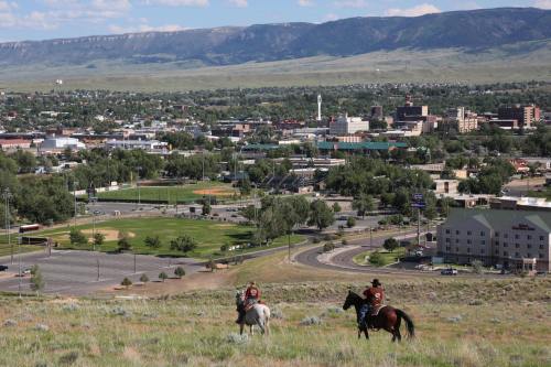 Two riders on horseback overlook a cityscape with green fields and mountains in the background.