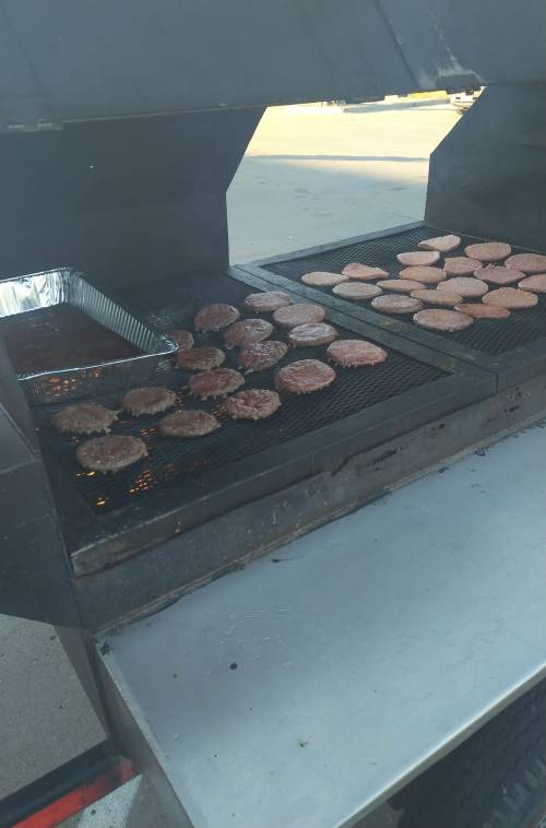A grill with multiple patties cooking on it, some in a tray, outdoors on a sunny day.