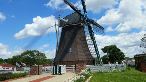 A large windmill with a brown roof and white blades, surrounded by green trees and a blue sky with fluffy clouds.