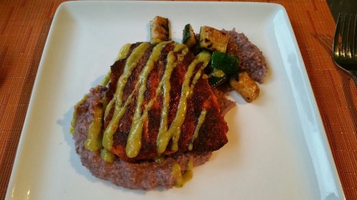 A plated dish featuring grilled fish with a mustard drizzle, served on a bed of purple mashed beans and grilled vegetables.