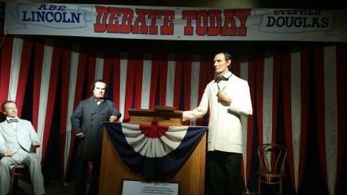 Wax figures of Abraham Lincoln and Stephen Douglas at a debate, with a patriotic backdrop and a podium.