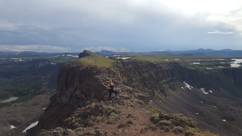 A person stands on a rocky cliff overlooking a vast mountainous landscape under a cloudy sky.