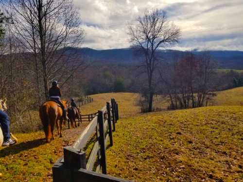 Two horseback riders on a trail, surrounded by autumn trees and rolling hills under a cloudy sky.