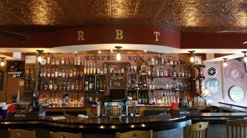 A well-stocked bar with various bottles on shelves, a wooden counter, and decorative ceiling tiles.