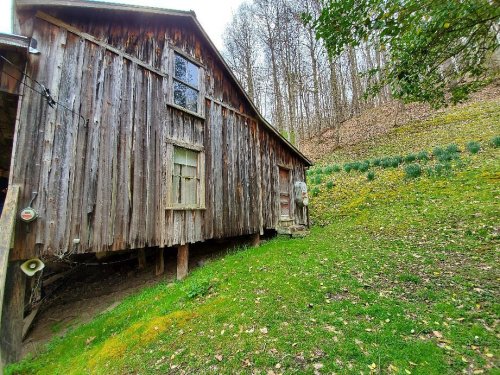 A weathered wooden barn stands on a grassy slope, surrounded by trees and wildflowers in a serene landscape.