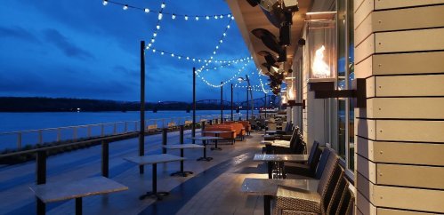 A riverside patio at dusk, featuring string lights, empty tables, and a view of the water and bridge in the distance.