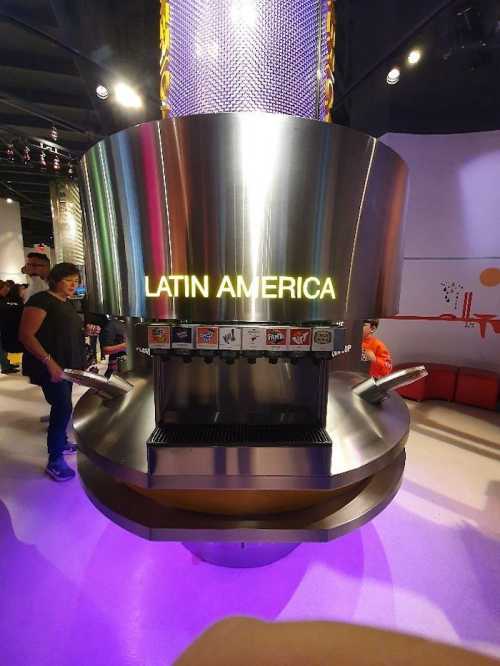 A modern beverage dispenser labeled "LATIN AMERICA" with various drink options, surrounded by visitors in a vibrant setting.