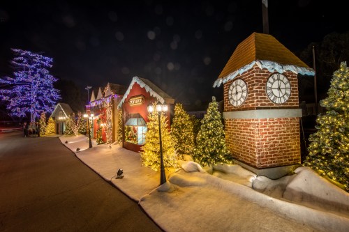 A festive street scene with gingerbread houses, illuminated trees, and a clock tower, all decorated for the holidays at night.