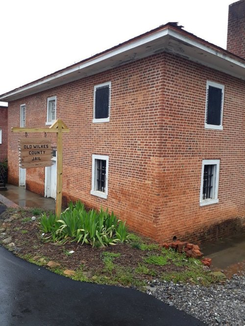 Historic brick building with barred windows, labeled "Old Wilkes County Jail," surrounded by greenery and gravel.