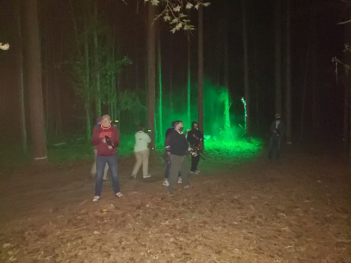 A group of people in a dark forest, illuminated by green light, with trees and fog in the background.