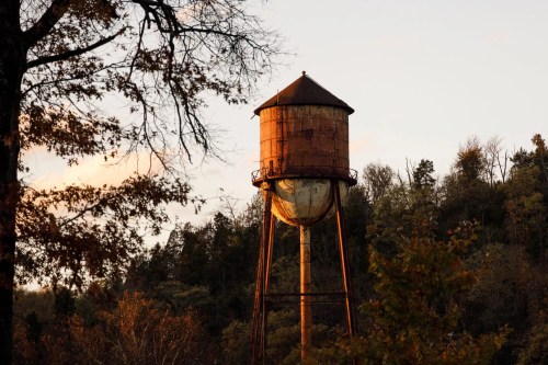 Rusty water tower at sunset, surrounded by trees and a warm golden light illuminating the scene.