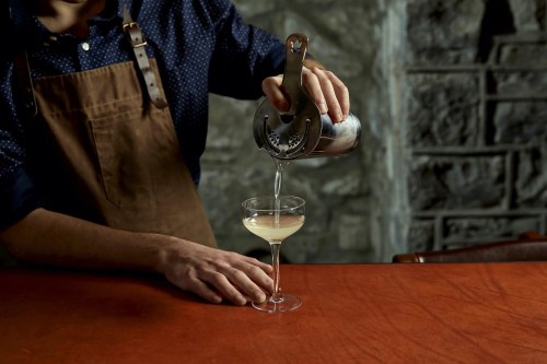 A bartender in an apron pours a cocktail from a shaker into a glass on a wooden bar.
