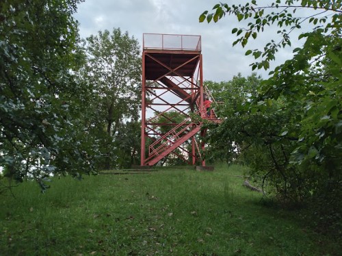 A red observation tower surrounded by greenery and trees under a cloudy sky.