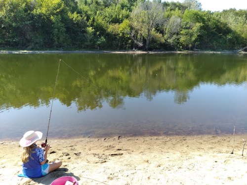 A child sits on the sandy bank of a river, fishing with a rod, surrounded by lush greenery and calm water.
