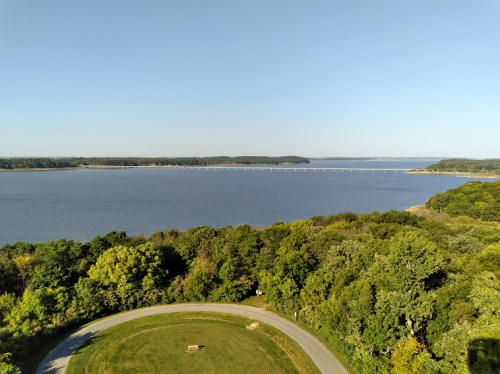 A scenic view of a calm lake surrounded by greenery, with a bridge visible in the distance under a clear blue sky.