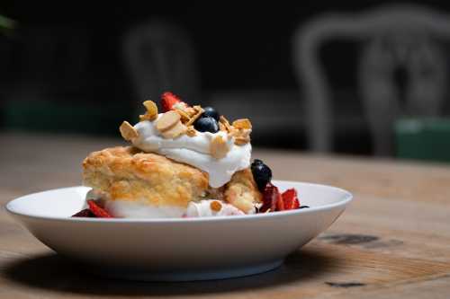 A delicious dessert featuring a biscuit topped with whipped cream, strawberries, blueberries, and granola in a white bowl.