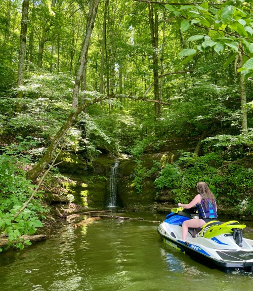 A person on a jet ski near a lush green forest with a small waterfall in the background.