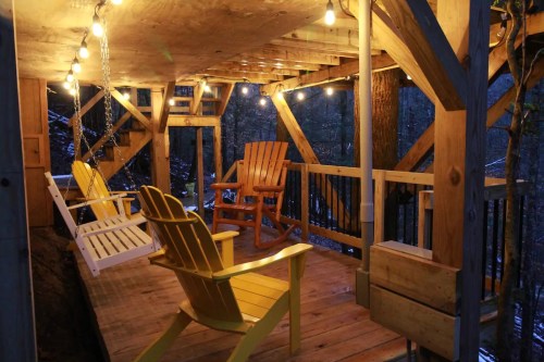 Cozy wooden deck with yellow and white chairs, illuminated by string lights, surrounded by trees at dusk.
