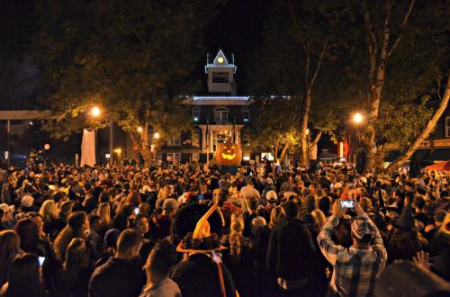 A large crowd gathers at night for a Halloween event, featuring a giant carved pumpkin in the center.