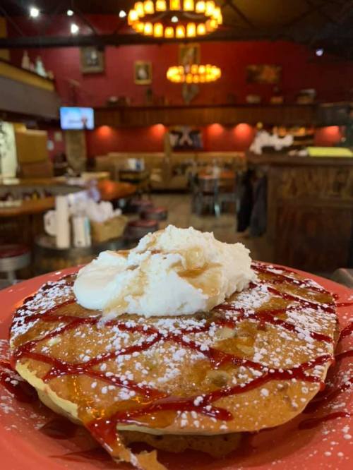 A close-up of a pancake topped with whipped cream and syrup, served on a red plate in a cozy restaurant setting.